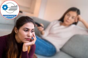 donna sul divano che guarda tv e un'altra triste con logo mediaset nel cerchio